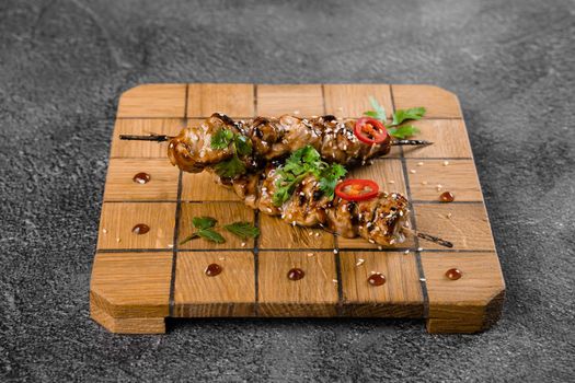 Meat on wooden skewers, garnished with cilantro, sesame, chili on square board on gray background