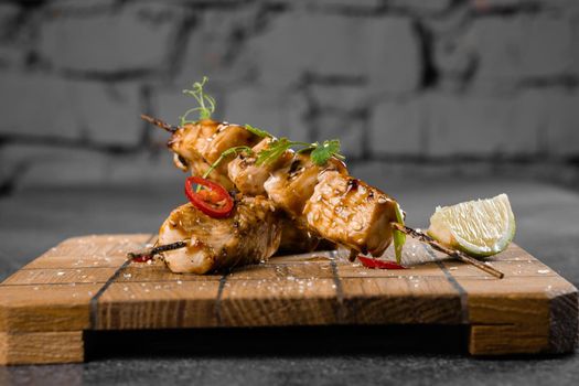 Meat on wooden skewers, garnished with cilantro, lime, sesame, chili on square board on gray background