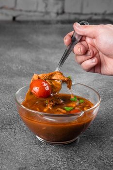 Soup with meat, mushrooms, tomatoes in transparent bowl on gray background. Appetizing thick satisfying tom yam