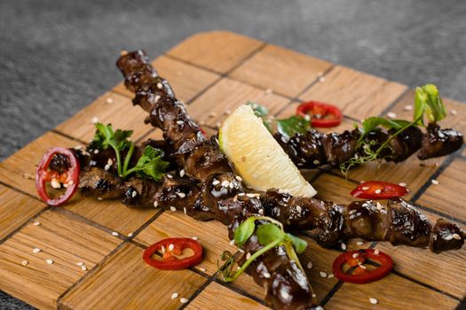 Grilled chicken hearts with lime on wooden skewer, garnished with chili, cilantro, sesame on gray background