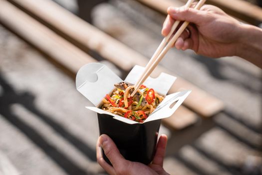 Wok in box in black food container. Holding spicy noodles with chopsticks. Fast food delivery service. Takeaway chinese street meal