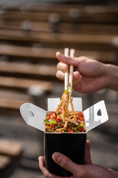 Wok in box in black food container. Holding spicy noodles with chopsticks. Fast food delivery service. Takeaway chinese street meal