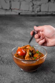 Soup with meat, mushrooms, tomatoes in transparent bowl on gray background. Appetizing thick satisfying tom yam