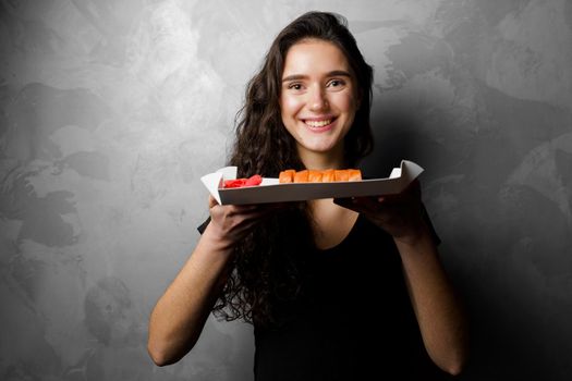 Girl holding philadelphia rolls in a paper box on gray background. Sushi, food delivery