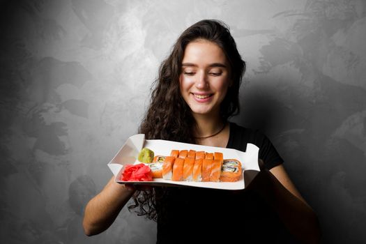 Girl holding philadelphia rolls in a paper box on gray background. Sushi, food delivery