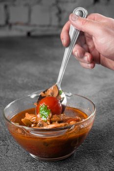 Soup with meat, mushrooms, tomatoes in transparent bowl on gray background. Appetizing thick satisfying tom yam