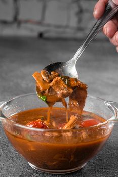 Soup with meat, mushrooms, tomatoes in transparent bowl on gray background. Appetizing thick satisfying tom yam