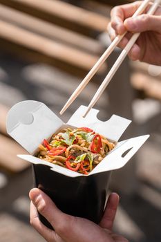 Wok in box in black food container. Holding spicy noodles with chopsticks. Fast food delivery service. Takeaway chinese street meal
