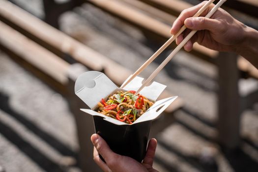 Wok in box in black food container. Holding spicy noodles with chopsticks. Fast food delivery service. Takeaway chinese street meal