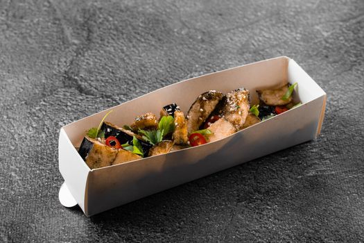 Fried eggplant dish in cardboard box. Food delivery. Vegetables garnished with cilantro and chili on gray background