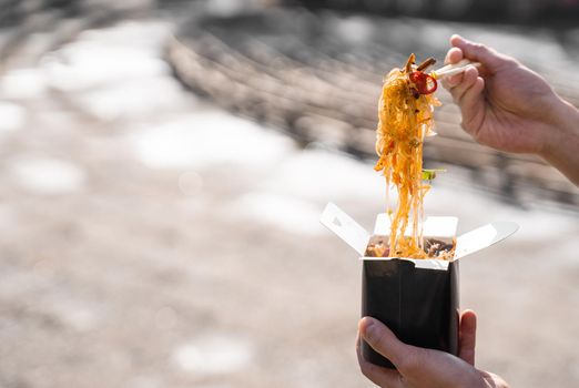 Wok in box in black food container. Holding rice noodles with chopsticks. Fast food delivery service. Takeaway chinese street meal. Eating udon.