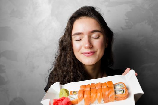 Surprised girl with sushi set philadelphia rolls in a paper box happy girl holding on a gray background. Food delivery