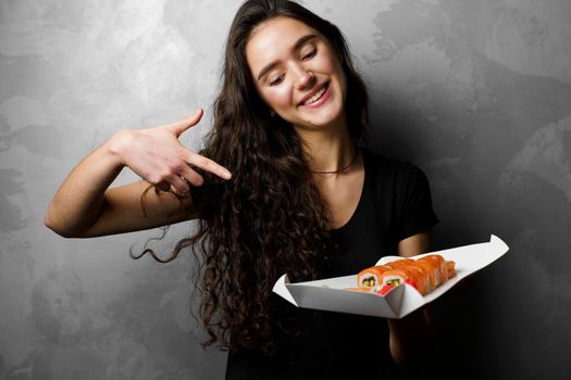 Surprised girl with sushi set philadelphia rolls in a paper box happy girl holding on a gray background. Food delivery