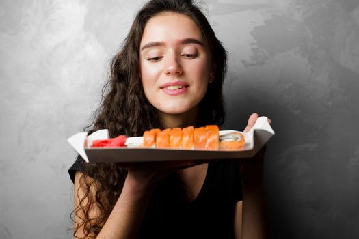 Surprised girl with sushi set philadelphia rolls in a paper box happy girl holding on a gray background. Food delivery