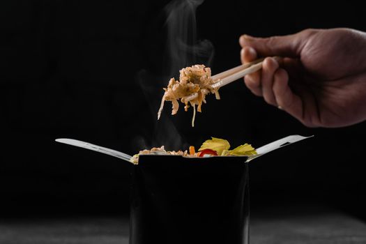 Steaming hot rice in wok box on black background. Holding rice with chinese chopsticks. Asian spicy dish for street restaurant and fast food delivery service