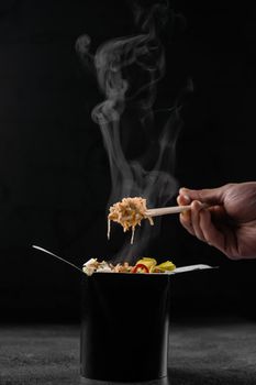 Steaming hot rice in wok box on black background. Holding rice with chinese chopsticks. Asian spicy dish for street restaurant and fast food delivery service