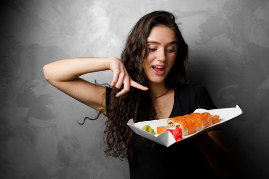 Surprised girl with sushi set philadelphia rolls in a paper box happy girl holding on a gray background. Food delivery