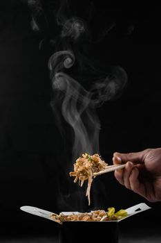 Steaming hot rice in wok box on black background. Holding rice with chinese chopsticks. Asian spicy dish for street restaurant and fast food delivery service