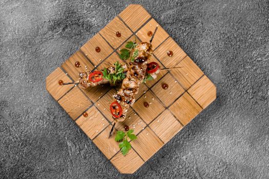 Meat on wooden skewers, garnished with cilantro, sesame, chili on square board on gray background