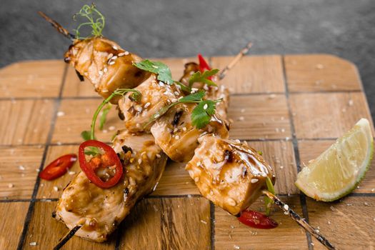 Meat on wooden skewers, garnished with cilantro, lime, sesame, chili on square board on gray background