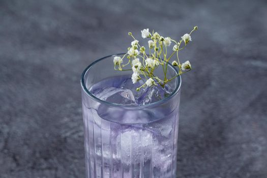 A cocktail in a tall glass with blue liqueur and ice, decorated with white flowers on gray background