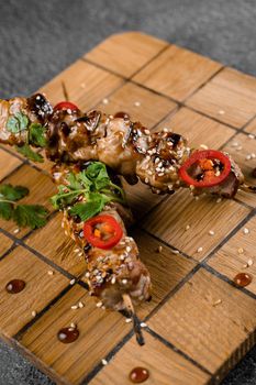 Meat on wooden skewers, garnished with cilantro, sesame, chili on square board on gray background