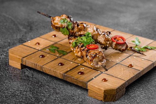 Meat on wooden skewers, garnished with cilantro, sesame, chili on square board on gray background