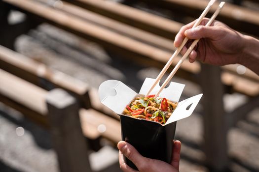 Wok in box in black food container. Holding spicy noodles with chopsticks. Fast food delivery service. Takeaway chinese street meal