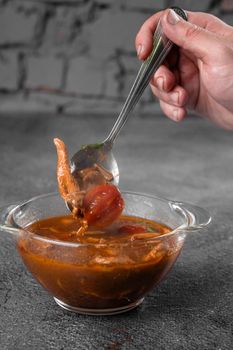 Soup with meat, mushrooms, tomatoes in transparent bowl on gray background. Appetizing thick satisfying tom yam