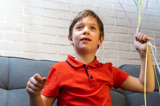 a happy European preschool boy is sitting on the sofa and smiling. The boy holds balloons inflated with helium by the ribbons. Birthday Celebration