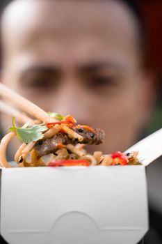 Chinese man holding spicy noodles with chopsticks. Wok in box in black food container. Fast food delivery service. Takeaway chinese street meal