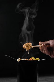 Steaming hot rice in wok box on black background. Holding rice with chinese chopsticks. Asian spicy dish for street restaurant and fast food delivery service