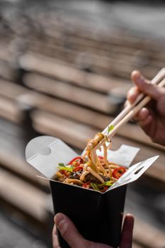 Wok in box in black food container. Holding spicy noodles with chopsticks. Fast food delivery service. Takeaway chinese street meal