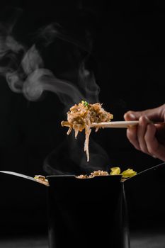 Steaming hot rice in wok box on black background. Holding rice with chinese chopsticks. Asian spicy dish for street restaurant and fast food delivery service