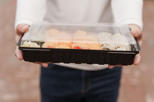 Closeup sushi in box healthy food delivery online service. Man in white clothes gives sushi set to you. Japanese cuisine: rolls, soy sauce, wasabi.