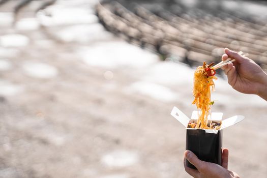 Wok in box in black food container. Holding rice noodles with chopsticks. Fast food delivery service. Takeaway chinese street meal. Eating udon.