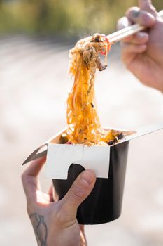 Wok in box in black food container. Holding rice noodles with chopsticks. Fast food delivery service. Takeaway chinese street meal. Eating udon.