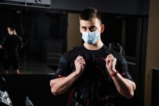 Man in EMS suit and medical mask in gym. Protection from coronavirus covid-19. Sport training in electrical muscle stimulation suit at quarantine period