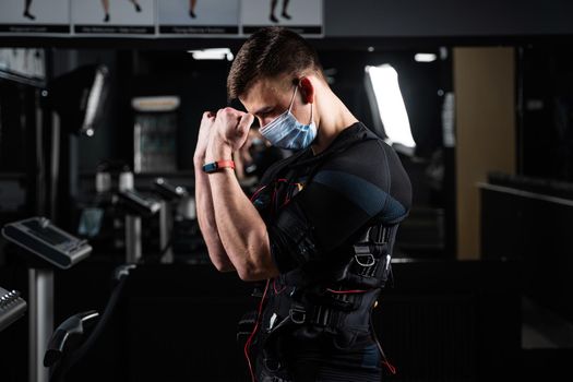 Man in EMS suit and medical mask in gym. Protection from coronavirus covid-19. Sport training in electrical muscle stimulation suit at quarantine period