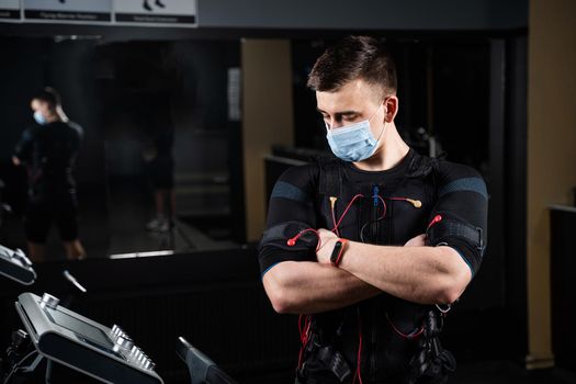 Man in EMS suit and medical mask in gym. Protection from coronavirus covid-19. Sport training in electrical muscle stimulation suit at quarantine period