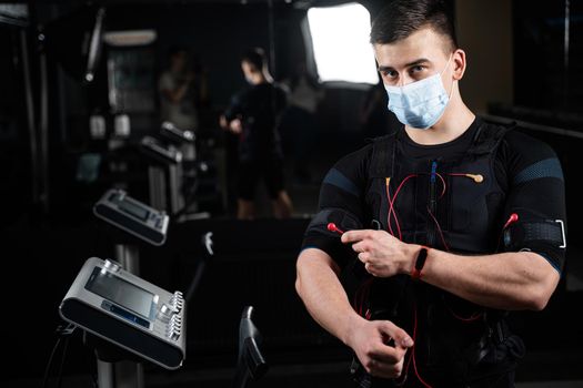 Man in EMS suit and medical mask in gym. Protection from coronavirus covid-19. Sport training in electrical muscle stimulation suit at quarantine period