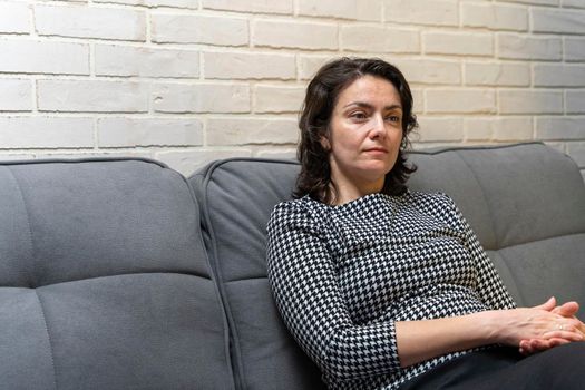 a middle-aged European woman with dark hair is sitting on a sofa against a light brick wall in a modern apartment. rest, thoughts