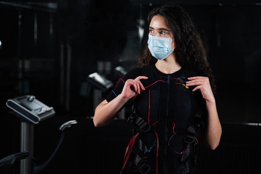 Girl in EMS suit and medical mask in gym. Protection from coronavirus covid-19. Sport training in electrical muscle stimulation suit at quarantine period