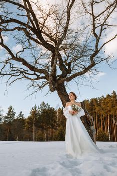 The bride is standing in a snow-covered forest next to a dry tree.