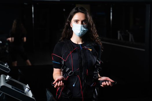 Girl in EMS suit and medical mask in gym. Protection from coronavirus covid-19. Sport training in electrical muscle stimulation suit at quarantine period