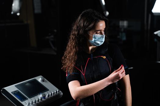 Girl in EMS suit and medical mask in gym. Protection from coronavirus covid-19. Sport training in electrical muscle stimulation suit at quarantine period