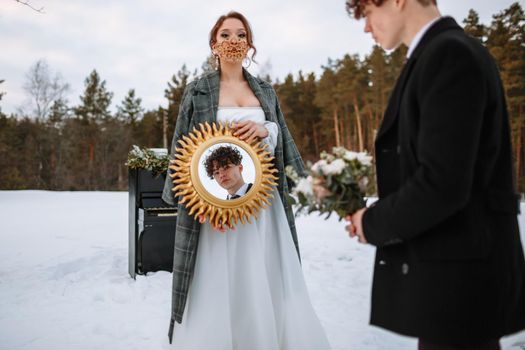 The groom looks in the mirror held by the bride. The bride is wearing a fabulous protective mask.