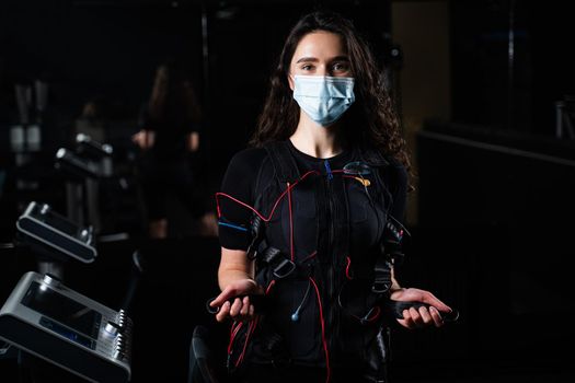 Girl in EMS suit and medical mask in gym. Protection from coronavirus covid-19. Sport training in electrical muscle stimulation suit at quarantine period