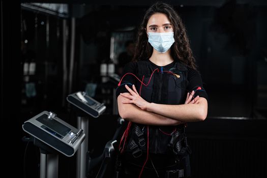 Girl in EMS suit and medical mask in gym. Protection from coronavirus covid-19. Sport training in electrical muscle stimulation suit at quarantine period