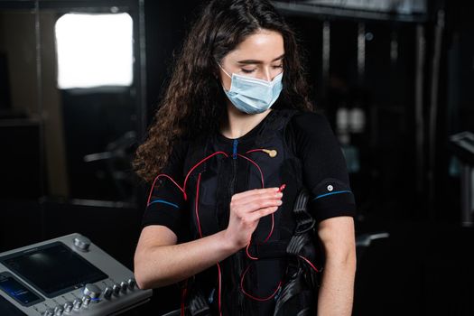 Girl in EMS suit and medical mask in gym. Protection from coronavirus covid-19. Sport training in electrical muscle stimulation suit at quarantine period